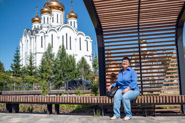 An elderly woman sits on a bench against the backdrop of a white Orthodox church with golden bathers.