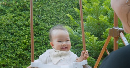 A happy baby enjoying a swing ride with a parent in a beautiful green garden. The baby is smiling and having fun on a bright sunny day.