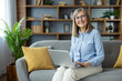 © Liubomir - Smiling senior woman working on laptop sitting on sofa. Comfortable home setting with bookshelves and plants in background. Concept of remote work, technology, senior lifestyle, happiness.
