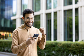 Wall Mural - Young man in glasses smiling and celebrating success while using smartphone outdoors. Business professional receiving good news, feeling happy. Modern office building background.