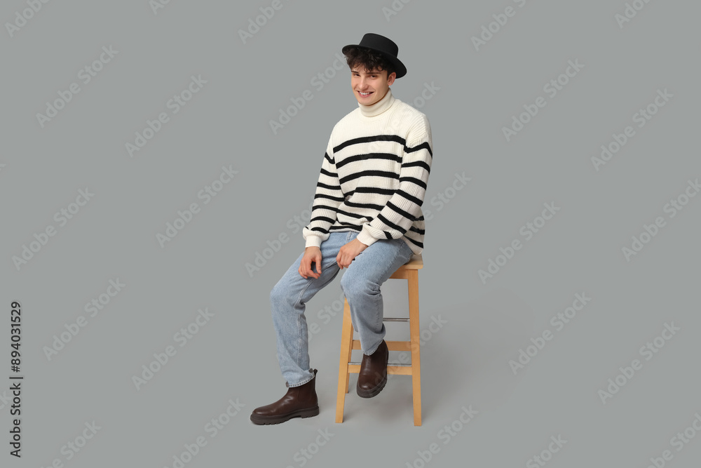 Handsome young man in stylish clothes and black hat sitting on stool against grey background