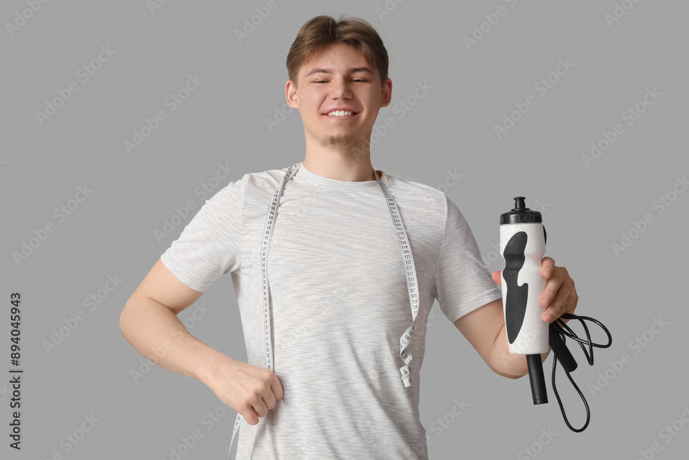 Sporty young man with bottle of water, jumping rope and measuring tape on grey background. Weight loss concept