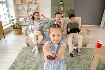 Wall Mural - Cute little girl with TV remote control and her family watching soccer game at home