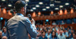 © Duka Mer - A businessman holding a microphone and giving a presentation to an audience in a conference hall, focusing on the back view of the man speaking to the crowd at a business meeting or town hall.