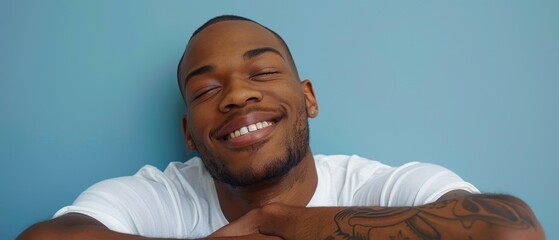 Close-up of a smiling man with tattoos, relaxing against a blue background, exuding positivity and happiness.