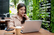 © bongkarn - A confident Asian businesswoman working remotely at a cafe, focusing on working on her laptop.
