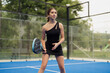 © arthurhidden - Young woman in black outfit playing padel tennis outdoors. She is focused and ready to hit the ball on a blue court.