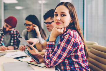 Fototapete - Portrait of positive attractive young woman designer dressed in casual wear holding smartphone and smiling at camera while sitting at meeting table with colleagues during working briefing in office
