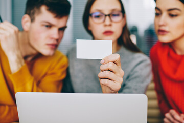 Fototapete - Pensive young people dressed in casual wear reading information on blank card sitting in office at netbook.Selective focus on business card with mock up area for your advertisement in woman's hand