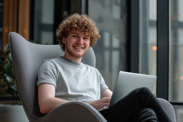 Young caucasian man with curly hair comfortably sitting in armchair working or using laptop with space for text
