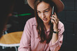 © BullRun - Wonder young woman in hat calling on smartphone and talking during phone conversation with client sitting at coffee table with notepad.Hipster girl in stylish hat explaining information on telephone