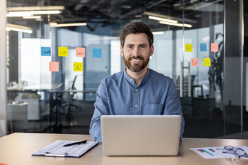 Portrait of a smiling young man working in a modern office using a laptop, sitting at a desk and looking at the camera