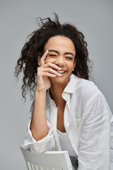 A young woman with curly hair smiles brightly while holding her hand to her face.