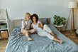 © Bliss - Two women relax in bed, sharing a cozy morning together, one sipping coffee while the other looks at her phone and smiles.