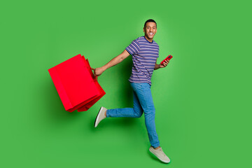 Sticker - Full body portrait of nice young man jump run smart phone shop bags wear t-shirt isolated on green color background