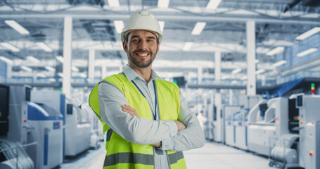 Poster - Portrait of Adult Caucasian Male Production Supervisor Looking at Camera And Smiling. Technician in a Hardhat And Reflective Jacket at an Industrial Factory with Heavy Machinery Producing Electronics.