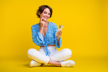 Poster - Full size photo of clever minded cute woman wear blue top sit look at smartphone finger on lips isolated on yellow color background