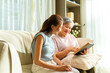 © CandyRetriever  - Asian senior mature woman and adult daughter spending time together reading a book on sofa in living room. Happy family relax and enjoy indoor lifestyle with leisure activity together at home.