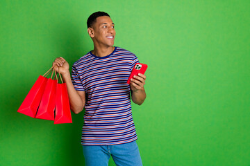 Poster - Photo of cheerful man wear striped stylish clothes hold packages purchase look empty space isolated on green color background