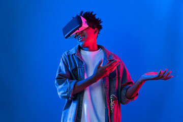 Young African American wearing VR pointing to empty space product launching brand explaining detail on blue pink neon light wall futuristic technology virtual reality at metaverse world. Contrivance.