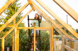 © rh2010 - Female carpenter using a power drill while working on a wooden greenhouse in her backyard. She is wearing casual clothes and work gloves, engaged in a DIY project, surrounded by nature