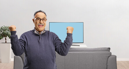 Wall Mural - Casual mature man with glasses gesturing happiness in front of tv