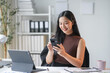 © amnaj - Asian businesswoman smiles as she works on her smartphone and laptop in a modern office, surrounded by paperwork. She exudes confidence and ambition in her bright workspace
