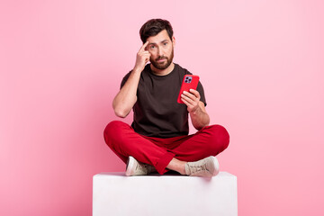 Poster - Full size photo of clever man dressed brown t-shirt sit on cube hold smartphone finger on forehead isolated on pink color background