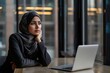 © David Zarzosa - Thoughtful businesswoman wearing a hijab, sitting at an office desk with a laptop.