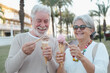 © luciano - Smiling retired couple having fun eating ice cream cone in the park. Joyful elderly lifestyle concept. Two white-haired seniors enjoying a sweet fresh dessert