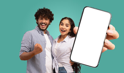 Fototapete - A young Indian couple stand against a teal background. They both look directly at the camera and smile. The woman holds a smartphone in front of them, displaying a blank white screen