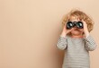 © panumas - Child in a striped shirt looking through binoculars with a beige background.