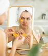 © JackF - Serene woman in forties in towel turban applying golden collagen eye patches in front of tabletop mirror, focusing on skin health in morning beauty ritual..