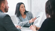 © Prostock-studio - A businesswoman is presenting information to two clients in an office setting. She is pointing at a document with a pen while sitting at a table with a tablet and other office supplies.