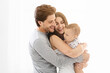 © Prostock-studio - This image shows a happy family of three, a man, a woman, and their baby, standing close together in a white interior setting. The man and woman are smiling at the baby