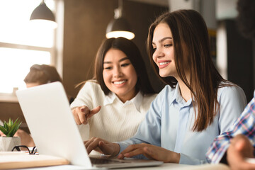 Aufkleber - Two multiethnic women are seated at a desk in a well-lit library, working together on a project. One is pointing at a laptop screen, suggesting an idea or solution