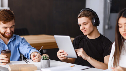 Poster - A group of young adults is gathered around a table in a well-lit office space, collaborating on a project. One is smiling while holding a digital tablet, another is taking notes