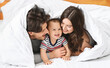 © Prostock-studio - This image shows a happy family of three in their bed. The mother, father, and baby are all smiling and looking at each other.
