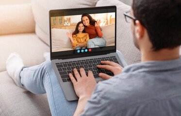 Poster - A man sits on a couch, wearing casual clothing and glasses, using a laptop to video chat with his wife and daughter. The woman and girl are sitting together, waving at the man.