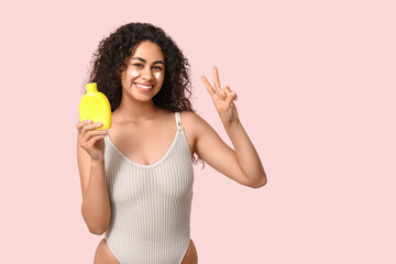 Wall Mural - Beautiful young happy African-American woman with bottle of sunscreen cream showing victory gesture on pink background