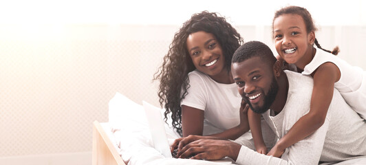 Wall Mural - Happy together. African american parents and their daughter lying on bed and using laptop, browsing internet or watching movie