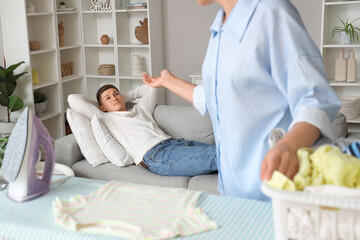 Wall Mural - Lazy husband lying on sofa while his wife ironing laundry at home