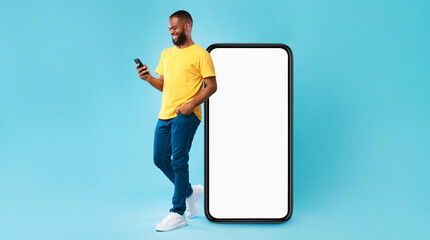 Handsome young Afro man using mobile phone while standing near huge smartphone with empty screen over blue studio background. Mockup for website or application, space for advertisement