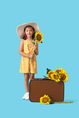 Cute little Asian girl with beautiful sunflowers and suitcase on blue background