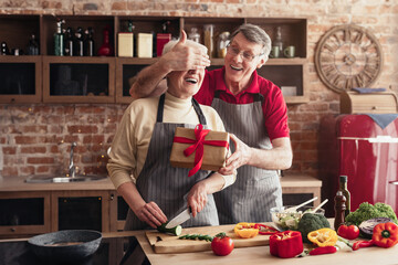 Wall Mural - Surprise. Happy senior woman receiving gift from her loving husband in kitchen, cooking together, empty space