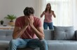 © MD Media - Young man sitting couch in front, crying and covering his face with hands while woman standing behind him is shouting at her husband. Domestic abuse concept