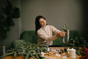 Wall Mural - Woman making Christmas wreath using natural pine branches, sitting near wooden table.