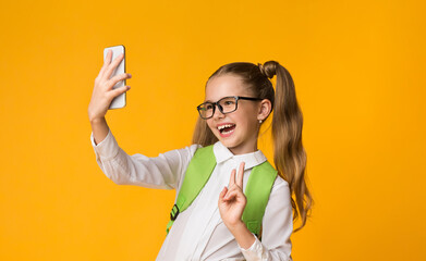 Wall Mural - School Selfie. Happy Schoolgirl Using Smartphone Taking Self-Portrait Gesturing Victory Sign On Yellow Studio Background. Copy Space