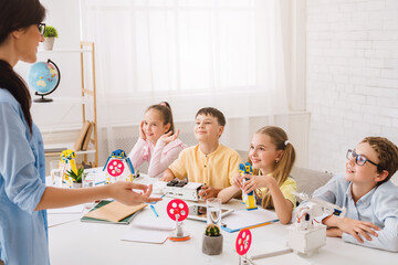 Poster - A young teacher instructs a group of elementary school students during a STEM activity. The students are sitting around a table, listening attentively to the teachers explanation.