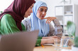 © Studio Marmellata - Two women in hijabs collaborate on a project in a modern office space One woman in a green hijab is smiling while the other in a blue hijab focuses on their work